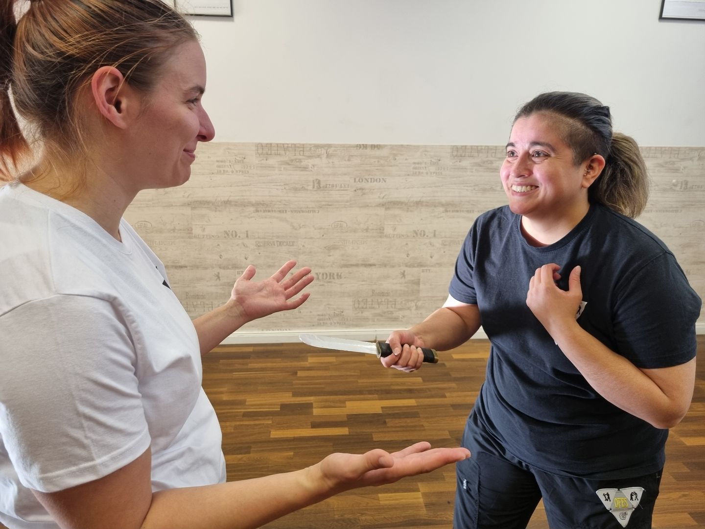 Zwei Frauen in Selbstverteidigungssituation, eine hält ein Trainingsmesser, beide stehen sich in geschützter Trainingsumgebung gegenüber