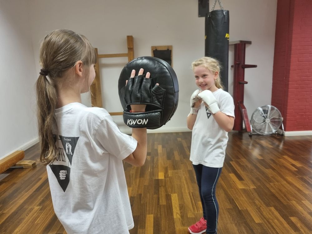 Zwei Mädchen im Wing Tsun Training, eines hält ein Schlagpolster, das andere trägt Boxhandschuhe und bereitet sich auf einen Schlag vor